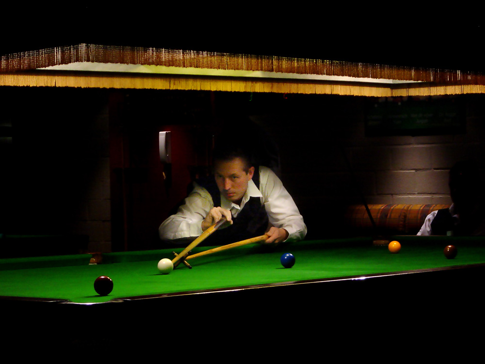 A snooker player using the rest to reach a shot across a full-size table.